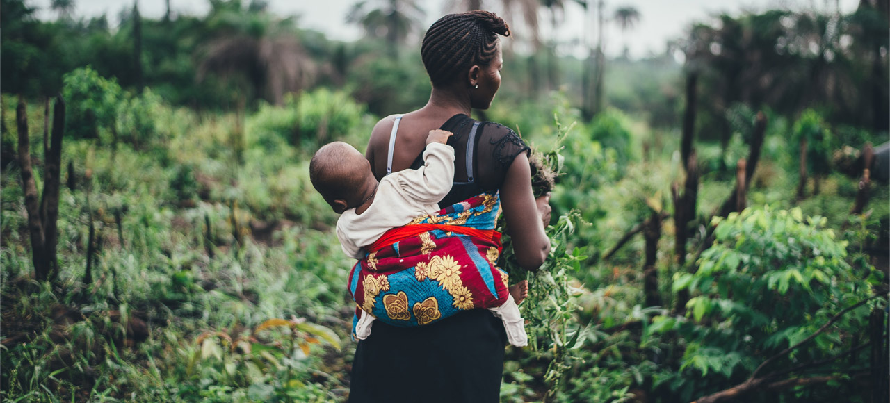 Mother carrying a young child on her back, shown from behind, in green fields in Sierra Leone. Credit: Annie Spratt, Unsplash.