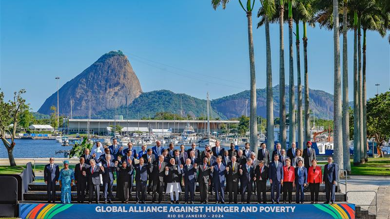 A group of people dressed in suits in front of a tropical background with sea, palm trees and mountains in the distance