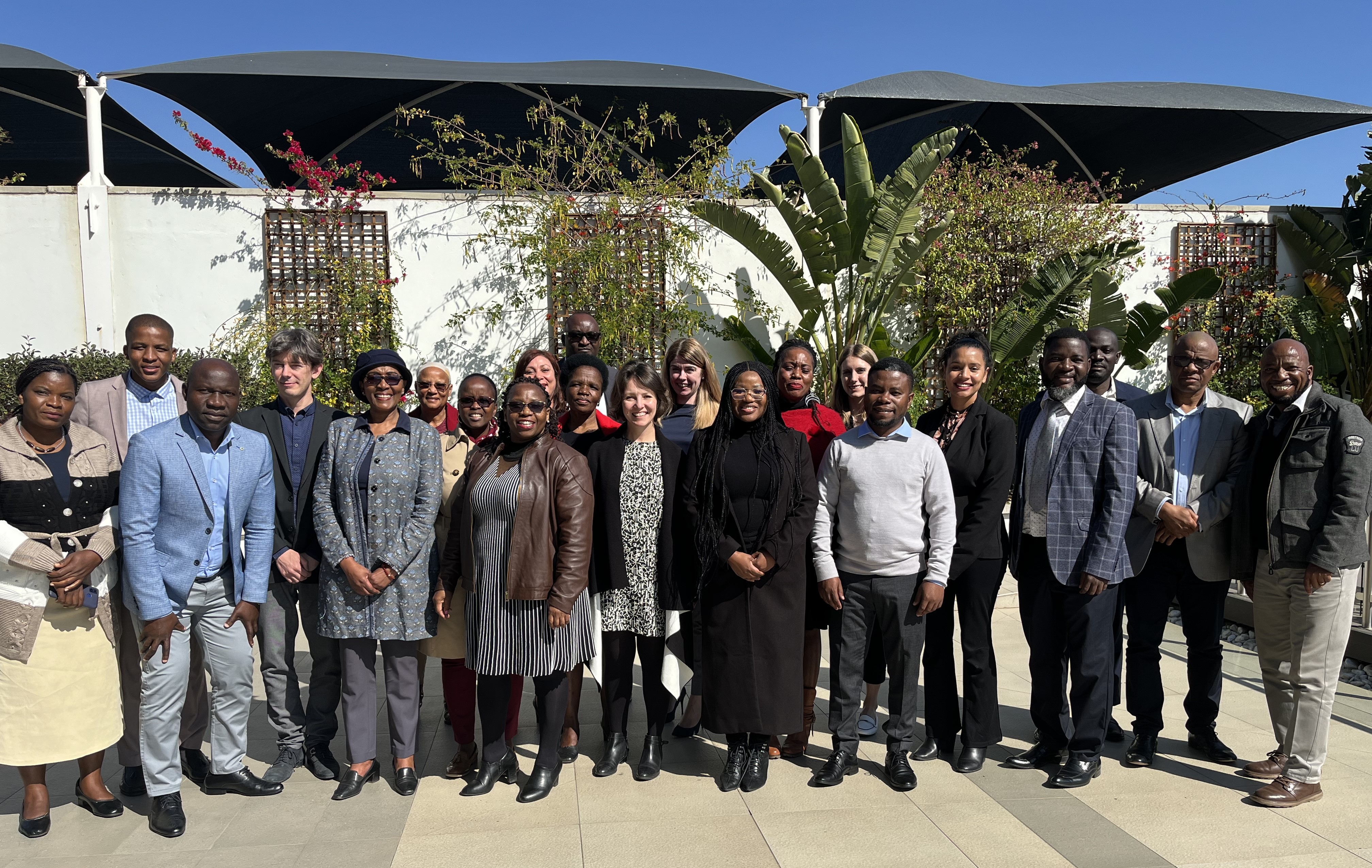 A group of about 20 people, both men and women, standing outdoors on a sunny day, posing together for a photo. They are dressed in a mix of professional and business-casual attire, with greenery and plants in the background.