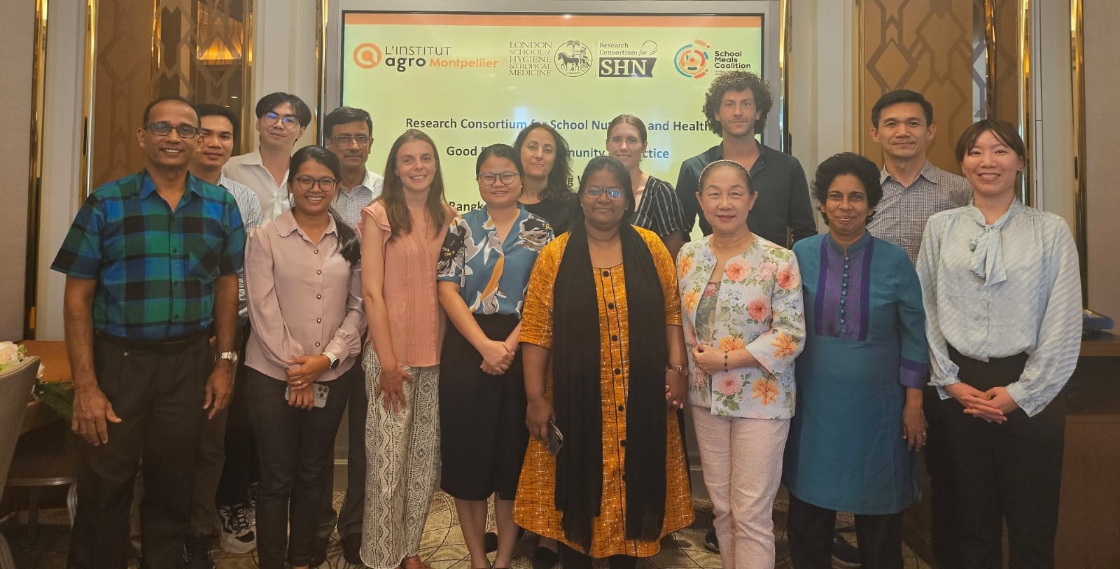 The image shows a diverse group of people standing indoors in front of a banner displaying logos from institutions like L&#039;Institut Agro Montpellier, LSHTM, and the School Meals Coalition. The banner mentions the &quot;Research Consortium for School Nutrition and Health&quot; and a &quot;Good Practices Community of Practice&quot; event in Bangkok. The group, dressed in casual and business-casual attire, is smiling for the camera, suggesting a collaborative workshop or conference focused on school nutrition and health.