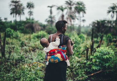 Mother carrying a young child on her back, shown from behind, in green fields in Sierra Leone