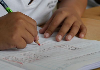 Image shows a child holding a red pencil, writing on paper