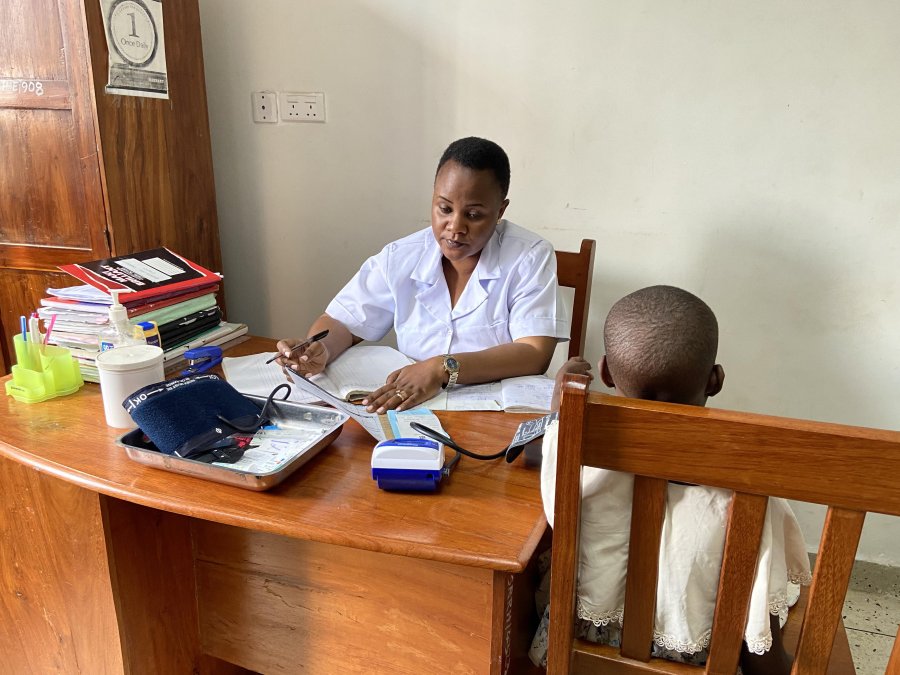 An African healthcare worker in a white uniform sits at a wooden desk, reviewing medical records with a young child patient.